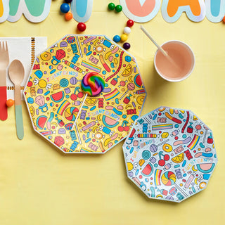 A colorful party setup featuring vibrant plates with candy illustrations, a wooden fork and spoon, beads, a pink cup, and a yellow background with cheerful decorations, creating a festive atmosphere.