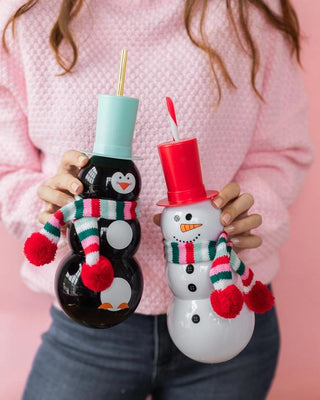A person holding two festive snowman bottles adorned with colorful scarves and hats against a pink background, evoking a cheerful holiday mood.