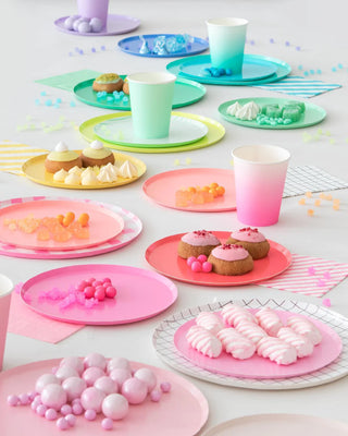A brightly colored dessert table setup with an assortment of pastel plates and cups, showcasing a variety of sweets such as cookies, candies, and meringues, arranged beautifully on a white surface for a festive celebration.