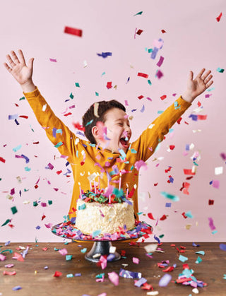A joyful child in a yellow shirt celebrates excitedly with arms raised high amid a shower of colorful confetti, standing behind a birthday cake on a pink background.