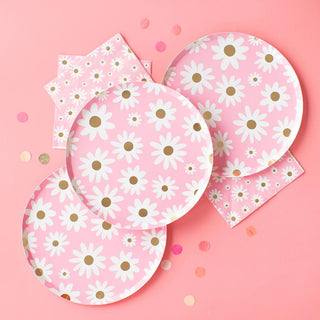 Three daisy-patterned paper plates are arranged on a pastel pink background, accompanied by scattered multicolored confetti, suggesting a cheerful, festive atmosphere, likely for a springtime or floral-themed party.