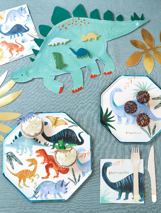 A dinosaur-themed party table setting featuring illustrated paper plates, a centerpiece with a cut-out dinosaur, and decorative foliage, accented with matching cups and utensils.