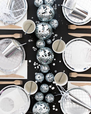 Table set for a party with metallic disposable plates, cups, and cutlery, adorned with several small disco balls scattered across a black tablecloth.