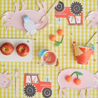 A whimsical children's party table setting with playful farm animal and tractor cutouts, vibrant colors, and fresh fruit on a picnic-style gingham cloth.