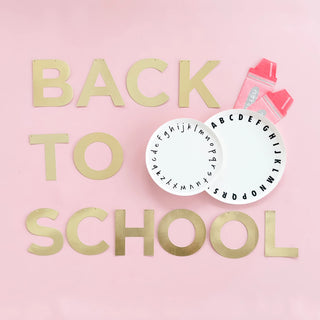 Golden letters spell "back to school" on a pink background alongside school supplies, including pencils, an eraser, and a circular paper alphabet chart, evoking a playful yet educational vibe.