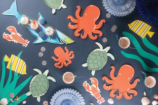 A colorful underwater-themed craft arrangement on dark background, featuring paper cutouts of fish, octopuses, turtles, and lobsters, complemented by decorative shells and pebbles.