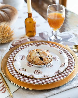 An elegant table setting featuring a golden plate with a checkered border, a baked pastry on a patterned dish, gold cutlery, and a glass of orange juice, evoking a warm and sophisticated brunch atmosphere.