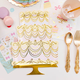A festive table setting featuring elegant, gold-patterned party plates, golden cutlery, pastel macarons, and whimsical party invitations, arranged on a white surface suggesting a sophisticated celebration.