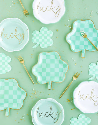 A festive st. patrick's day table setting featuring plates with "lucky" inscribed, clover-shaped napkins, golden cutlery, and scattered confetti on a green backdrop.