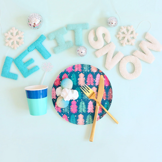 Festive scene with a paper cup, plate of cake pops, and gold cutlery on a blue background. Felt letters spell "LET IT SNOW" with snowflake and ornament decorations.