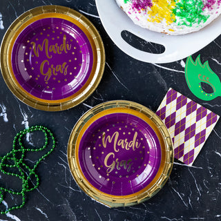 Mardi gras celebration setup with themed purple plates reading "mardi gras," green beads, a festive mask, and a decorated cake on a marble background.