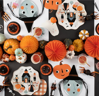 A festive halloween table setup with themed paper plates, napkins, and decorations including pumpkins, paper fans, and whimsical ghost and witch illustrations on a black backdrop.