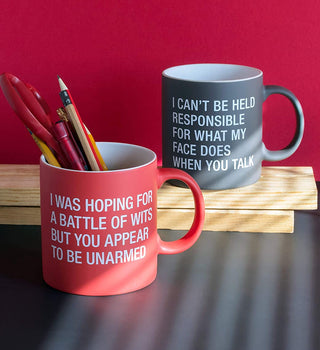 Two humorous mugs on a desk; the red mug, holding pens, reads "i was hoping for a battle of wits but you appear unarmed," and the gray mug says "i can't be held responsible for what my face does when you talk.