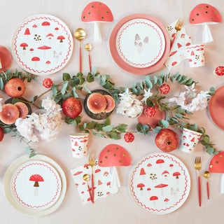 Table set with mushroom-themed plates, cups, napkins, gold utensils, pomegranates, and eucalyptus leaves, decorated with flowers and standing mushroom ornaments.