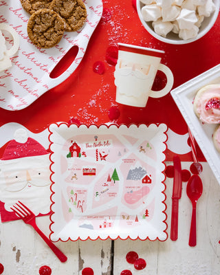 A festive holiday place setting featuring a whimsical north pole-themed placemat, red cutlery, a santa mug, cookies, and meringues on a white and red speckled backdrop.