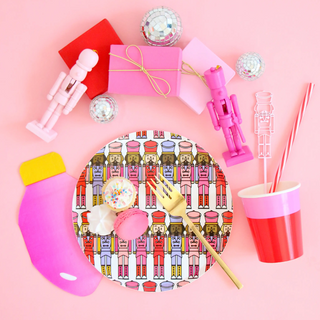 A vibrant birthday party setup with a toy soldier-themed plate, colorful envelopes, disco balls, plastic cutlery, a cup, and a whistle, all on a pink background.