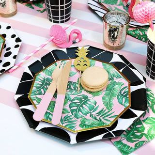 A tropical-themed party table setting with a hexagonal plate featuring a leaf pattern, a macaron, wooden cutlery, and pineapple decoration.