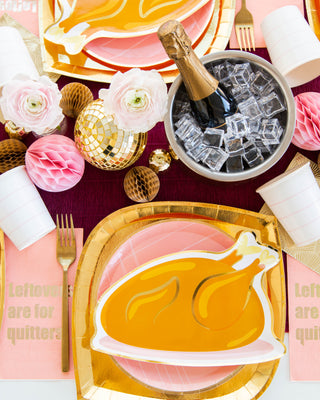 A festive table setting with gold and pink decorations, featuring paper plates with turkey illustrations, pink napkins reading "Leftovers are for quitters," and a champagne bottle in an ice bucket.