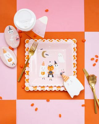 A whimsical halloween-themed flat lay with a decorated plate, golden cutlery, and themed cookies, all set against a vibrant orange and pink background, accented by scattered candies.