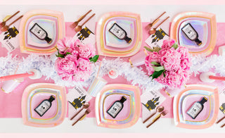 A pastel-themed party table setting with hexagonal plates, pink tablecloth, decorative peonies, and party favors, exuding a soft, celebratory atmosphere.