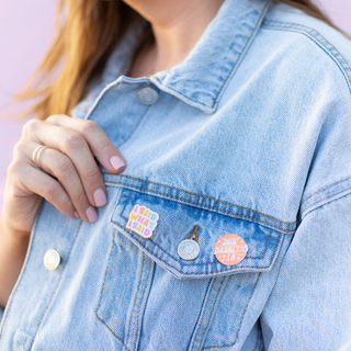 A person sporting a light blue denim jacket adorned with colorful enamel pins on the lapel, set against a soft pink background.
