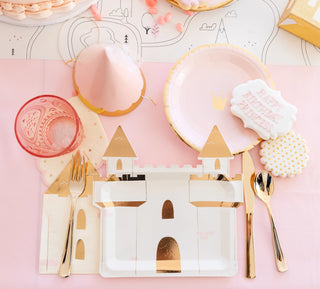 A festive table setting featuring a castle-themed party plate, pastel pink and gold utensils, a crown-adorned napkin, and delicately decorated cookies, all laid upon a soft pink tablecloth for a whimsical birthday celebration.