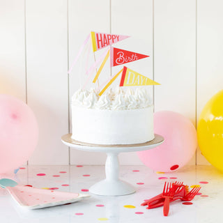 A festive birthday cake with white frosting stands on a pedestal, adorned with colorful "happy birthday" banners, surrounded by balloons and confetti, signaling a cheerful celebration.