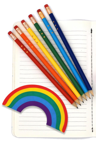 Seven colored pencils, each a different color of the rainbow, arranged together on an open lined notebook. A semi-circle rainbow eraser is placed beside them.