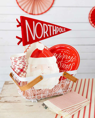 Basket with red and white holiday-themed items, including a North Pole flag, Santa plates, striped cups, and napkins on a wooden surface.