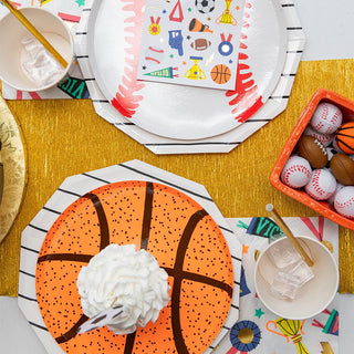 A vibrant table setup featuring sports-themed party plates, a basketball-designed plate topped with a swirl of whipped cream, a red basket with ball toys, and colorful napkins; all set upon a gold runner, indicating a festive sports-themed celebration.