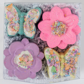An assortment of colorful, flower and bunny-shaped bath bombs decorated with glitter and sprinkles, presented in a gift box with crinkle paper.