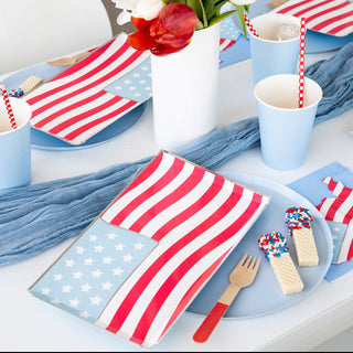 A festive display with paper fan decorations in red, white, and blue, themed for an american patriotic celebration, featuring star motifs, a garland, and a dessert table with cake and beverages.