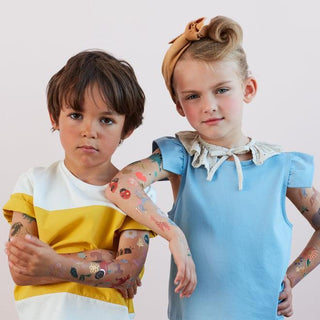 Two children showing off their temporary tattoos with confidence, the boy in a striped yellow shirt, and the girl in a blue dress with a bow in her hair.
