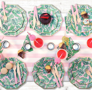 A vibrant summer table setting with tropical-patterned plates, pink utensils, and palm leaf napkins, adorned with colorful cupcakes, macarons, and refreshing beverages for a festive outdoor gathering.