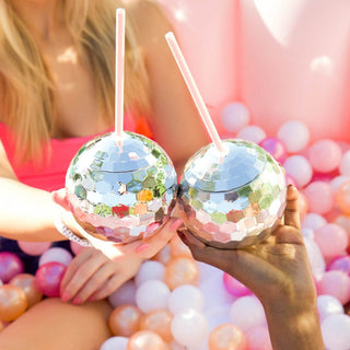 Two people hold disco ball-shaped sippy cups with pink straws against a backdrop of pastel balloons, symbolizing a festive and glamorous celebration.