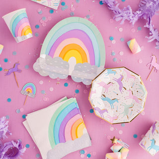 A vibrant party setup features pastel rainbow-themed decorations, including paper plates, napkins, cups, and confetti, with unicorn and rainbow motifs scattered across a pink backdrop.