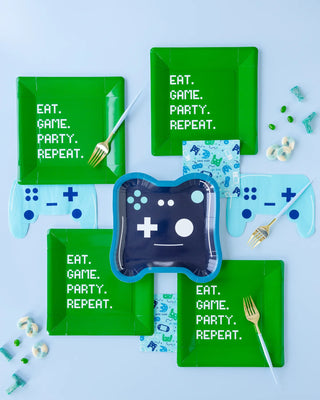 Top-down view of a gaming-themed party setup with green plates that read "EAT. GAME. PARTY. REPEAT.", blue game controller-shaped napkins, blue utensils, and a controller-shaped plate in the center.