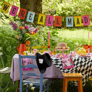 A whimsical outdoor tea party setup featuring a table with colorful decorations, flowers, a teapot, and a cake. A banner reading "We're All Mad" is hung above the table.