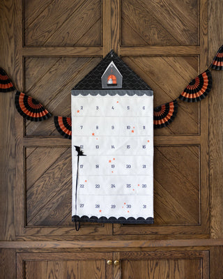 The Countdown to Halloween Canvas Pocket Advent Calendar by My Mind’s Eye, featuring 31 numbered pockets, hangs on a wooden door, flanked by black and orange fan decorations. Perfect for a spooky countdown to Halloween!