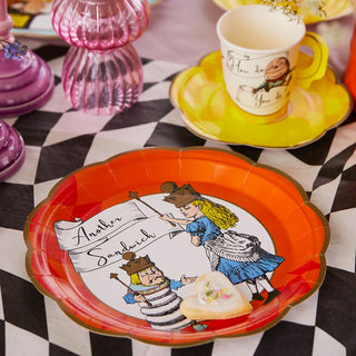 Close-up of a table setting featuring a red decorative plate from the Talking Tables' Alice in Wonderland Bright Scalloped Paper Plates collection with an illustration and the text "Another Sandwich," a yellow teacup on a matching saucer, and a heart-shaped cookie.