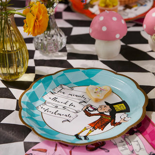 The Alice in Wonderland Bright Scalloped Paper Plates by Talking Tables, featuring classic Alice illustrations and the text "Ten minutes allowed for refreshments," sit on a checkered tablecloth. Surrounded by flower vases and mushroom figurines, they make a charming addition to any Alice collection.