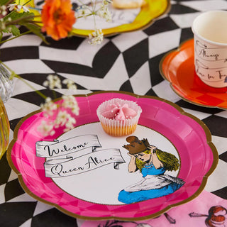 A decorated table set with a pink Alice in Wonderland Bright Scalloped Paper Plate by Talking Tables featuring classic drawings of Alice as a queen, "Welcome Queen Alice" text, a cupcake, and additional themed tableware on a checkered tablecloth from the enchanting Alice collection.