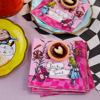 A stack of pink napkins featuring classic Alice in Wonderland illustrations saying "Let's be Seated" is set next to Talking Tables' Alice in Wonderland Bright Scalloped Paper Plates and a small tart with a heart-shaped topping.