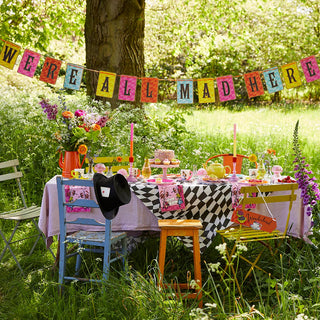 Outdoor tea party setup with a checkered tablecloth, colorful decorations, Talking Tables' Alice in Wonderland Bright Scalloped Paper Plates featuring classic Alice illustrations, a black top hat on a chair, and a “WE'RE ALL MAD HERE” banner hung between trees in a grassy, sunlit area.