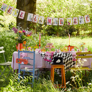 A colorful outdoor tea party scene with whimsical decorations, flowers, and a "We're All Mad Here" banner. Various chairs and table settings from the Alice collection adorn the space amidst lush greenery, each place showcasing classic Talking Tables' Alice in Wonderland Bright Scalloped Paper Plates.