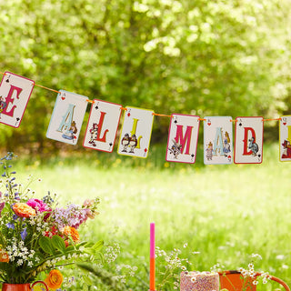 The vibrant Alice in Wonderland Multi Colored Double Sided Bunting from Talking Tables, featuring letters and illustrations spelling "ALL MAD HERE," is strung outdoors. Below the bunting, a table adorned with colorful flowers and whimsical tea party decorations is set. This setup creates the perfect atmosphere for any Alice in Wonderland-themed gathering.