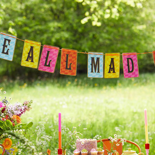 In a lush, green outdoor setting adorned with flowers and candles on the table below, the Talking Tables' Alice in Wonderland Multi Colored Double Sided Bunting spells "WE'RE ALL MAD," evoking an enchanting Alice in Wonderland tea party atmosphere.