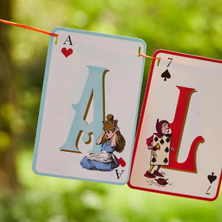 Two playing cards hanging by an orange string: the left card is the Ace of Hearts with a character in a blue dress, and the right card is the Seven of Spades with a character in red attire. This whimsical double-sided bunting from Talking Tables evokes an Alice in Wonderland tea party vibe and is named "Alice in Wonderland Multi Colored Double Sided Bunting.