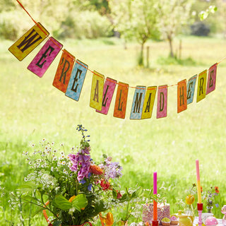 An Alice in Wonderland Multi Colored Double Sided Bunting from Talking Tables with the message "WE'RE ALL MAD HERE" hangs above an outdoor tea party table setting adorned with flowers, candles, and assorted decorations.