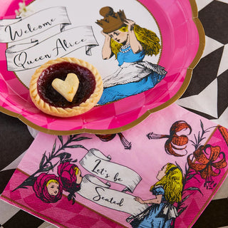 A heart-shaped tart on a pink plate with an illustration of a girl labeled "Queen Alice." A matching fairy tale napkin from the Alice in Wonderland Pink Paper Napkins collection by Talking Tables, featuring another illustration of the girl and the text "Let's be Seated," evokes an enchanting tea party setting.